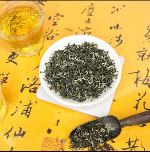 Loose leaf Jasmine Snowflake Tea displayed in a white bowl on a traditional Chinese calligraphy mat, with a wooden scoop and a glass of brewed tea beside it.