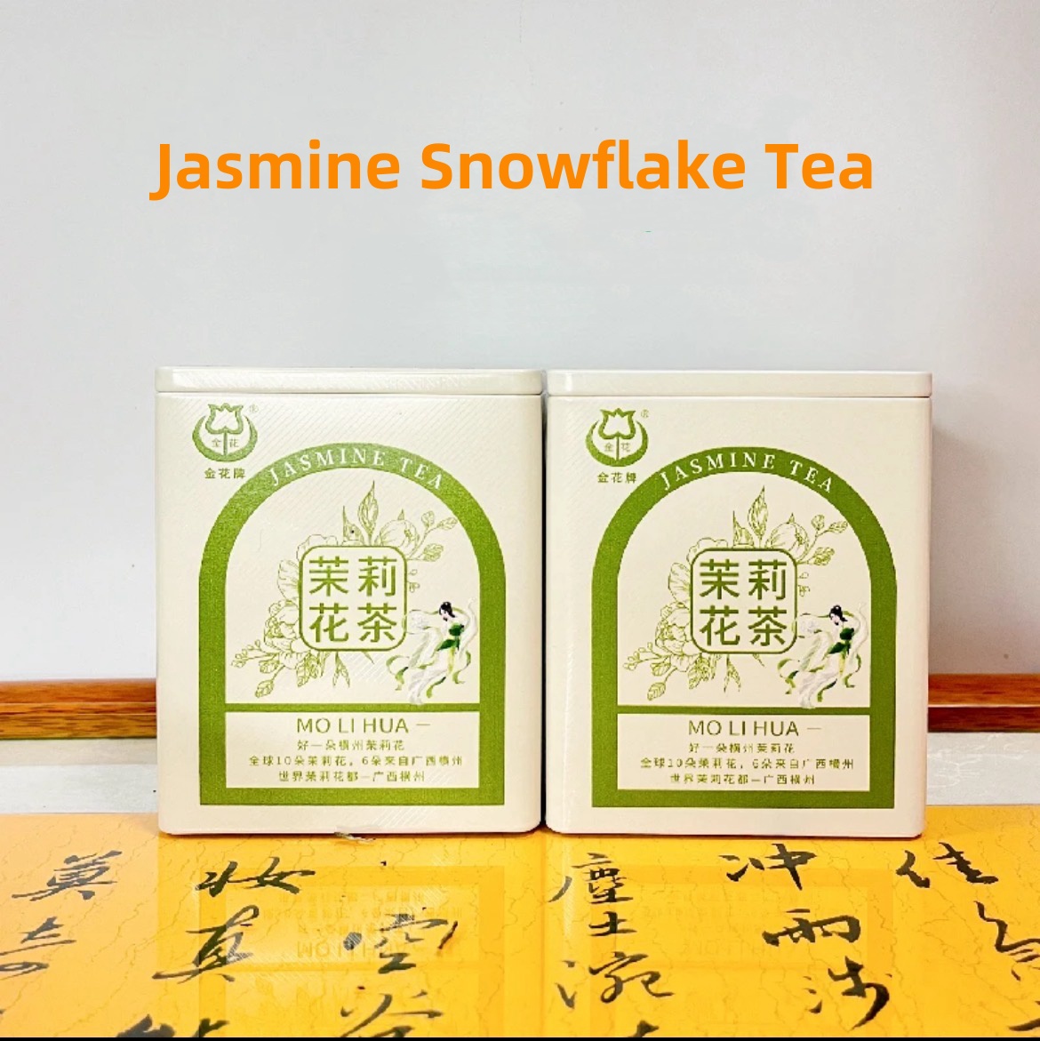 Two tins of Jasmine Snowflake Tea placed on a yellow calligraphy mat, featuring traditional Chinese packaging and floral green tea branding